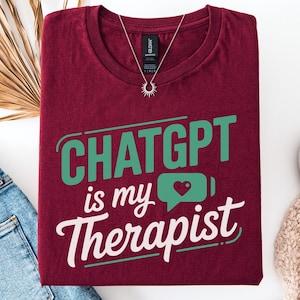 Peut inclure: T-shirt bordeaux avec l'inscription "ChatGPT is my Therapist" en vert et blanc. Le motif comprend une bulle de dialogue avec un cœur et une icône de batterie. Un collier argenté et des chaussures blanches sont également visibles.