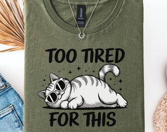 Puede incluir: Camiseta verde oliva con la frase "TOO TIRED FOR THIS" en negro. Debajo, un gato de dibujos animados con gafas de sol. La camiseta se muestra doblada con vaqueros azules y zapatos blancos.