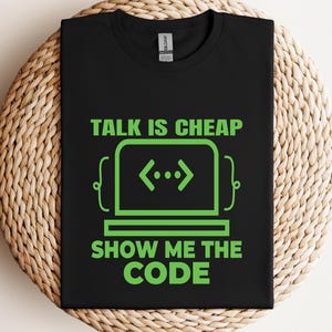 May include: Black t-shirt with the text "TALK IS CHEAP SHOW ME THE CODE" in bright green. The design includes a stylized laptop with code symbols. The shirt is displayed on a woven surface.