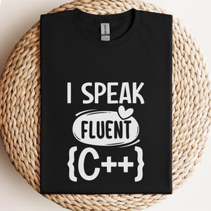 Könnte beinhalten: Schwarzes T-Shirt mit dem weißen Text "I Speak Fluent C++". Der Text ist zentriert, mit "Fluent" in einem Oval mit einem Herz darüber. Das T-Shirt wird auf einer gewebten Oberfläche präsentiert.