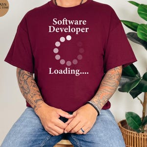 May include: Maroon t-shirt with the text "Software Developer Loading...." in white. The shirt has short sleeves and a crew neck. The image also includes a logo that says "The Print Studio" and "3-5 DAYS SHIPPING TIME".
