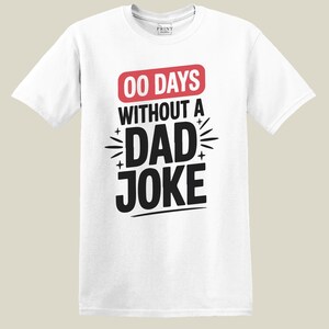 May include: White t-shirt with the text "00 DAYS WITHOUT A DAD JOKE" in black and red. The shirt has a crew neck and short sleeves. The text is centered on the front of the shirt.