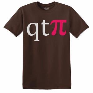 Peut inclure: T-shirt marron avec le texte "qtπ" en blanc et rose. Le t-shirt a des manches courtes et un col rond. Le logo The Print Studio est visible en haut à gauche.