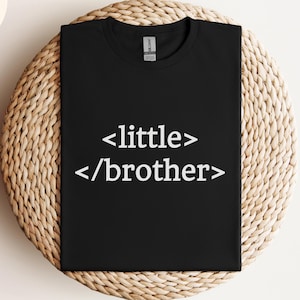 Puede incluir: Camiseta negra con el texto "<little> /brother>" en blanco. La camiseta se muestra sobre una superficie circular tejida. Un círculo negro con la palabra "BLACK" está en la esquina superior derecha. El logotipo de la marca y el texto "WORLDWIDE SHIPPING" están en la esquina superior izquierda.