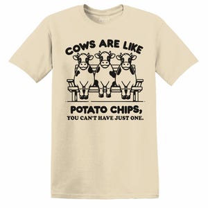Puede incluir: Camiseta color arena con un gráfico negro. El gráfico presenta tres vacas de dibujos animados sentadas en un banco, con el texto "COWS ARE LIKE POTATO CHIPS, YOU CAN'T HAVE JUST ONE." La camiseta tiene mangas cortas y cuello redondo.