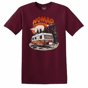 May include: Maroon t-shirt featuring a graphic of a vintage camper with the text "NOMAD at HEART" in orange and white. The design includes a sunset, trees, and a mountain range. The shirt is a short-sleeved crew neck.