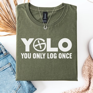 May include: A military green t-shirt with the text "YOLO YOU ONLY LOG ONCE" in white. The shirt is laid flat with a pair of blue jeans, white sneakers, and a necklace. The shirt is a casual wear item.