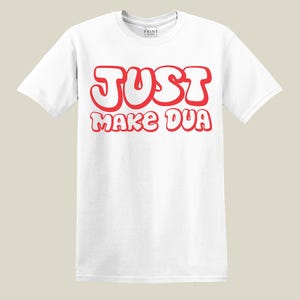 May include: White t-shirt with the text "JUST MAKE DUA" in red, bubble-style letters. The shirt has short sleeves and a crew neck. The Print Studio logo is visible in the upper left corner.