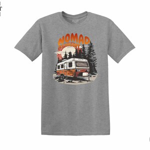May include: A heather grey t-shirt featuring a graphic of a vintage camper in a forest scene with the text "NOMAD ADVENTURE". The design includes a sunset and pine trees, creating a nature-inspired theme.