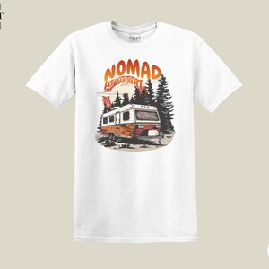 May include: White t-shirt featuring a graphic of a vintage camper with the text "NOMAD AT HEART" in orange and red. The design includes trees and a sunset, evoking a sense of adventure and travel.
