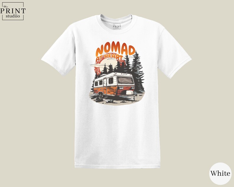 May include: White t-shirt featuring a graphic of a vintage camper with the text "NOMAD AT HEART" in orange and red. The design includes trees and a sunset, evoking a sense of adventure and travel.