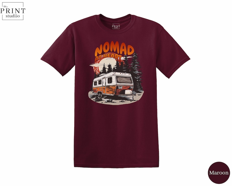 May include: Maroon t-shirt featuring a graphic of a vintage camper with the text "NOMAD at HEART" in orange and white. The design includes a sunset, trees, and a mountain range. The shirt is a short-sleeved crew neck.