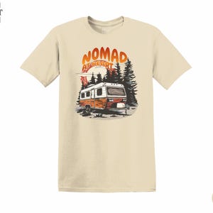May include: Sand-colored t-shirt featuring a graphic of a vintage camper in a forest setting. The design includes the text "Nomad at Heart" in orange and white. The shirt is made of a soft material.