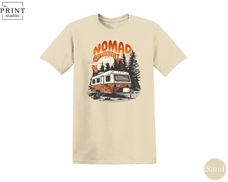 May include: Sand-colored t-shirt featuring a graphic of a vintage camper in a forest setting. The design includes the text "Nomad at Heart" in orange and white. The shirt is made of a soft material.