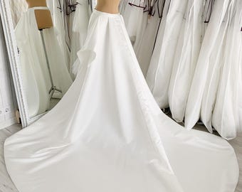 Puede incluir: Una falda de novia de satén blanco con una superposición de volantes y una larga cola. La falda se muestra en un maniquí en un entorno de tienda.