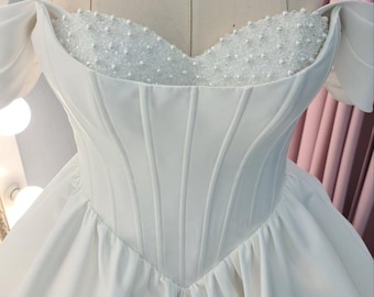 Könnte beinhalten: Ein weißes, schulterfreies Brautkleid mit herzförmigem Ausschnitt und einem perlenbesetzten Oberteil. Der Rock ist voll und fließend.