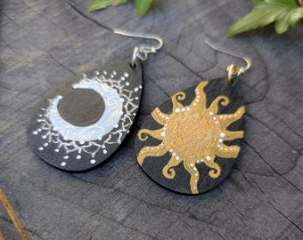 May include: A pair of teardrop-shaped earrings. One earring features a white crescent moon and star design on a black background. The other earring has a gold sun design with white accents on a black background. The earrings have silver hooks.