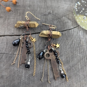 May include: A pair of dangle earrings featuring horizontal raw brass-toned crystals wrapped in copper wire. Each earring includes a skeleton key charm, black faceted crystal beads, thin gold-toned chains, and a small gold-toned star charm.