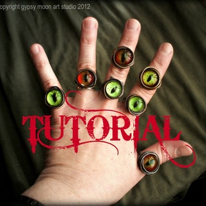 May include: A hand wearing several rings with green and orange eye-shaped stones. The rings are made of metal and have a vintage look. The word "TUTORIAL" is written in red across the hand.