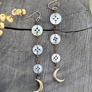 May include: A pair of dangle earrings featuring three white, four-hole buttons connected by copper-colored wire. Each earring terminates in a small, metallic crescent moon charm suspended from the bottom button, attached to a gold-toned ear wire hook.