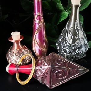 May include: A collection of decorative bottles and stoppers in different shapes and colours. The bottles are metallic, in shades of red, silver, and bronze. One bottle has a skull design. The stoppers are white.