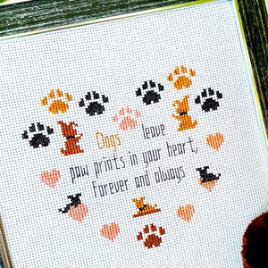 May include: Framed cross-stitch art with a black and gray frame. The artwork features paw prints, dog silhouettes, and hearts in shades of brown, black, and peach. Text reads "Dogs leave paw prints in your heart, forever and always."