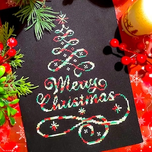 May include: A black canvas with a cross-stitched Christmas tree design in red, green, and white. The words "Merry Christmas" are stitched below the tree. The canvas is surrounded by Christmas decorations.