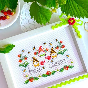 May include: A white framed cross-stitch embroidery with a bee and strawberry theme. The embroidery features three bees, three strawberries, and the text "bee sweet".