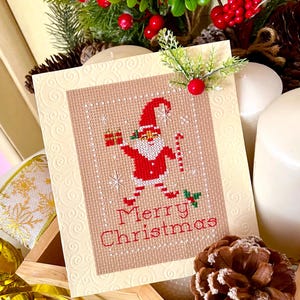 May include: A cream-colored greeting card with an embossed swirl pattern featuring a cross-stitch design of a Santa Claus figure holding a gift and a candy cane. The text Merry Christmas is stitched in red below the figure, accented by a small sprig.