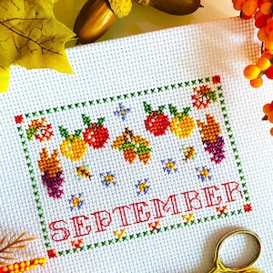 May include: Cross-stitch embroidery with a seasonal theme. The design features the word "SEPTEMBER" in red, surrounded by images of apples, grapes, and leaves in autumn colors. Gold scissors and fall foliage are in the background.