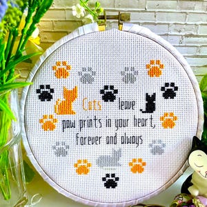 以下が含まれることがあります： 「Cats leave paw prints in your heart, forever and always」の文字が入ったクロスステッチのフープアート。オレンジ、黒、グレーの足跡と猫のシルエットのデザイン。刺繍糸と装飾的なハサミが含まれています。