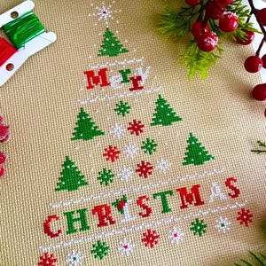 May include: A cross-stitch design featuring a Christmas tree shape with the word "CHRISTMAS" in red and green. The design includes green trees, snowflakes, and the words "Merry" in red and green. The background is a tan fabric.