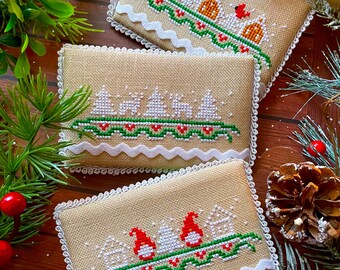 Puede incluir: Tres adornos navideños rectangulares de punto de cruz beige con bordes festoneados blancos. Cada uno presenta un diseño festivo diferente: casas, árboles y gnomos, todos con acentos rojos, verdes y blancos. Los adornos están dispuestos sobre una superficie de madera con vegetación festiva.