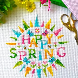 Puede incluir: Un diseño de punto de cruz sobre tela blanca que dice "HAPPY SPRING" en letras coloridas. El diseño incluye mariposas, flores y rayos de sol en tonos de rosa, azul, verde y amarillo. Tijeras doradas y otros suministros de artesanía en el fondo.