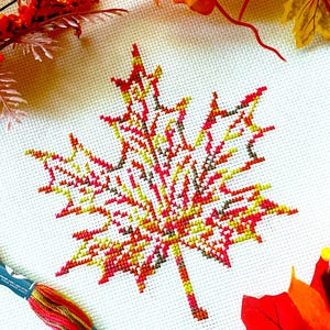 May include: A cross-stitch maple leaf in autumn colors of red, yellow, and brown. The leaf is centered on a white fabric background, with a skein of embroidery floss and decorative fall foliage in the corners.