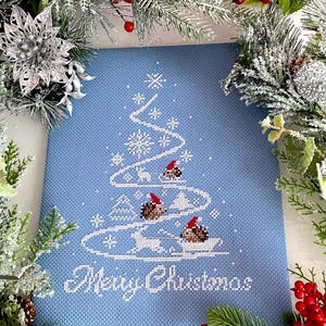 Op de afbeelding: Een blauw kruissteekontwerp van een kerstboom met egels die kerstmanmutsen dragen. De tekst "Merry Christmas" is onder de boom geborduurd.