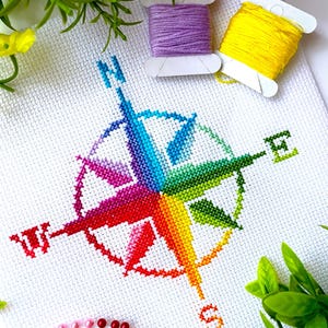 Könnte beinhalten: Ein fertiggestelltes Kreuzstichprojekt mit einem bunten Kompass-Design. Die Kompassrose ist in Regenbogenfarben auf weißem Stoff gestickt. Spulen mit lila und gelbem Garn sind zu sehen, zusammen mit Nadeln und Grün.