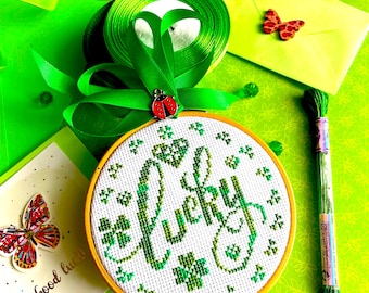 Könnte beinhalten: Ein grüner und weißer Stickrahmen mit dem Wort "lucky", das mit grünem Garn gestickt ist. Der Rahmen ist von grünem Band, einem grünen Umschlag und einer Karte mit der Aufschrift "viel Glück" umgeben.