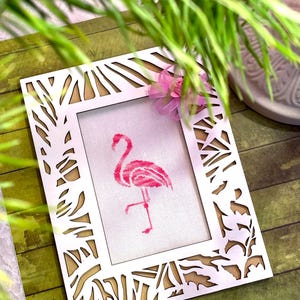 May include: A white wooden picture frame with a cutout design of leaves and branches. The frame holds a pink cross-stitch of a flamingo.