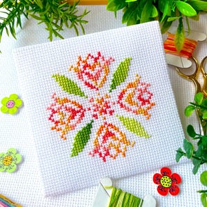 May include: A close-up of a cross-stitch embroidery featuring a design of five orange and pink tulips with green leaves. The embroidery is on white Aida cloth and is surrounded by green and orange embroidery floss.