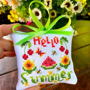 May include: A square white cross-stitch fabric ornament featuring the text Hello Summer in red and green. The design includes embroidered red cherries, a slice of watermelon, yellow sunflowers, and small bees. A bright lime green ribbon is tied at the top.