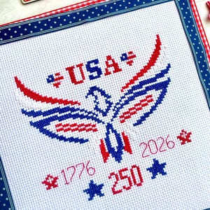 May include: A patriotic cross-stitch design featuring a red, white, and blue eagle emblem with the word "USA" and the numbers "1776" and "250". The design is framed with a blue and white star ribbon and a red and white gingham ribbon.