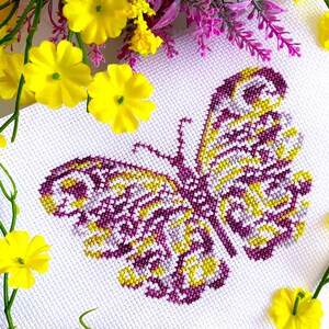 May include: A purple and yellow butterfly cross-stitch pattern on white Aida cloth. The butterfly is surrounded by yellow flowers.
