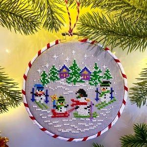 Puede incluir: Un adorno navideño de punto de cruz que representa una escena invernal con muñecos de nieve esquiando y en trineo. La tela gris está enmarcada por un borde a rayas rojas y blancas. El adorno está suspendido por una cuerda roja y blanca.