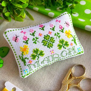 Puede incluir: Una almohada blanca bordada a punto de cruz con un borde verde, bordada con flores, tréboles y la palabra "MARCH" en verde. Una mariquita y una mariposa también están bordadas en la almohada. Tijeras doradas e hilo en primer plano.