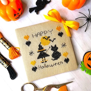May include: A cross-stitch piece with the words "Happy Halloween" and a witch and cat design. The image includes orange pumpkins, a black spider, and gold scissors. The piece is decorated with yellow and black accents.