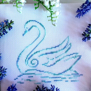 May include: A blue and green cross-stitch embroidery of a swan on a white background. The swan is in a swimming pose with its neck curved and its head turned to the side. The embroidery is surrounded by sprigs of white and purple flowers.