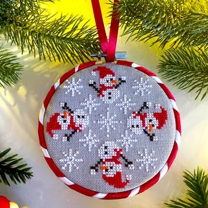 May include: A cross-stitched Christmas ornament featuring four snowmen in red hats and scarves, surrounded by snowflakes on a gray background. The ornament is framed in a red and white striped border and has a red ribbon for hanging.