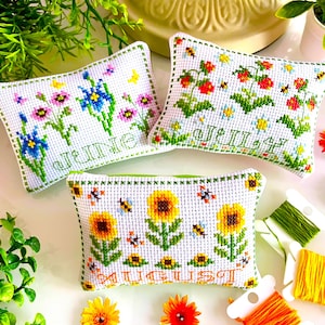 May include: Three white cross-stitch pillows with floral designs and month names: June, July, and August. The pillows feature colorful flowers, bees, and sunflowers. Green and orange embroidery thread and gold scissors are also visible.