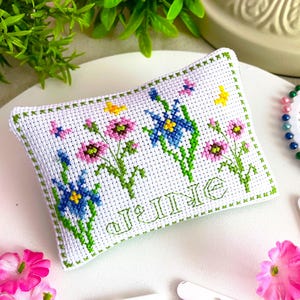 May include: A white cross-stitch pillow with a floral design featuring blue, pink, and yellow flowers, and butterflies. The word "JUNE" is stitched in green. The pillow is surrounded by green foliage and pink flowers.